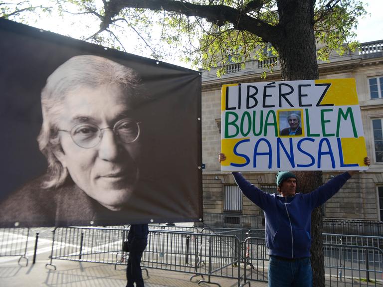 Auf der Straße protestiert ein Mann mit einem Banner für die Freilassung von Boualem Sansal, links daneben ein Plakat, das den franko-algerischen Schriftsteller zeigt.