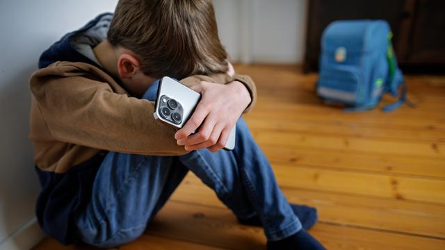 Ein Junge sitzt mit einem Smart-Phone in der Hand neben seinem Schul-Ranzen und versteckt seinen Kopf. 