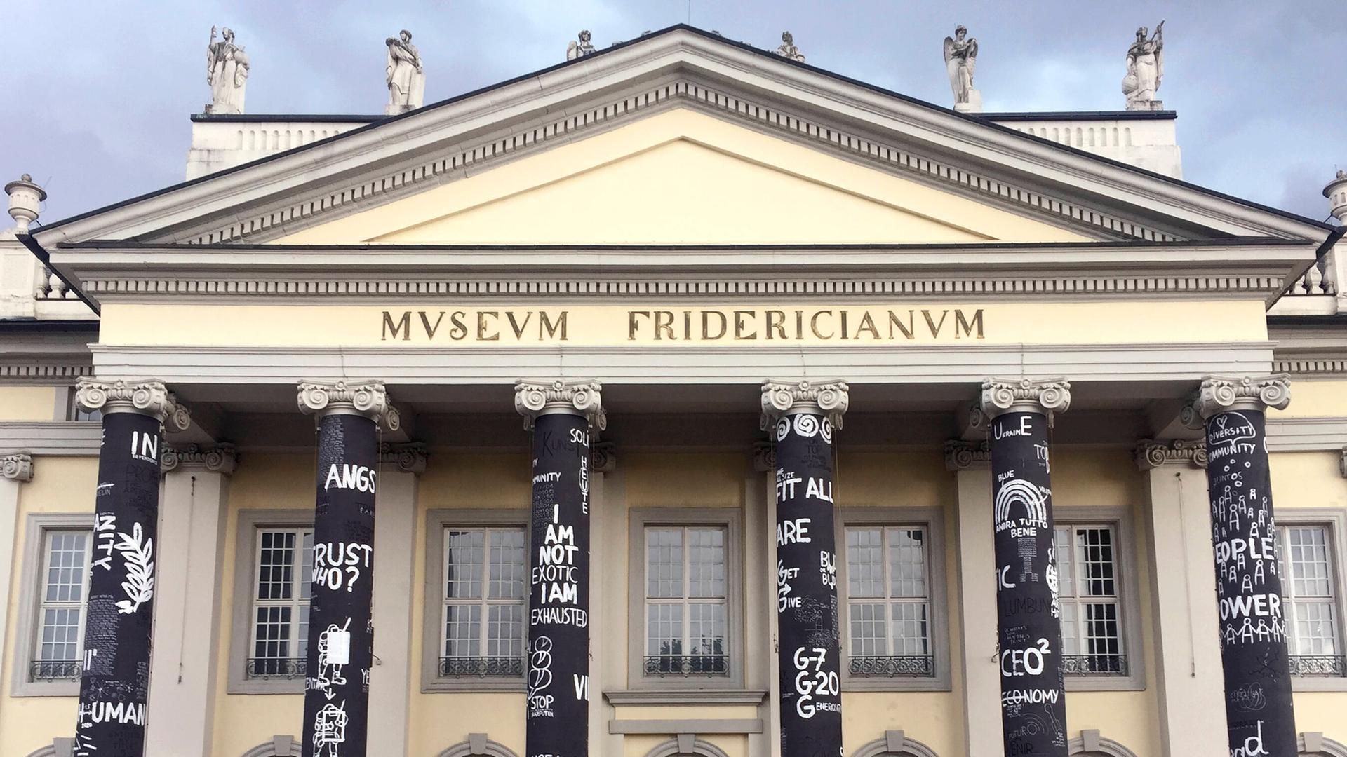 Der Eingang von dem Museum Fridericianum. 6 Eingangs-Säulen sind mit Plakaten geschmückt. Vor dem Eingang stehen viele Menschen.