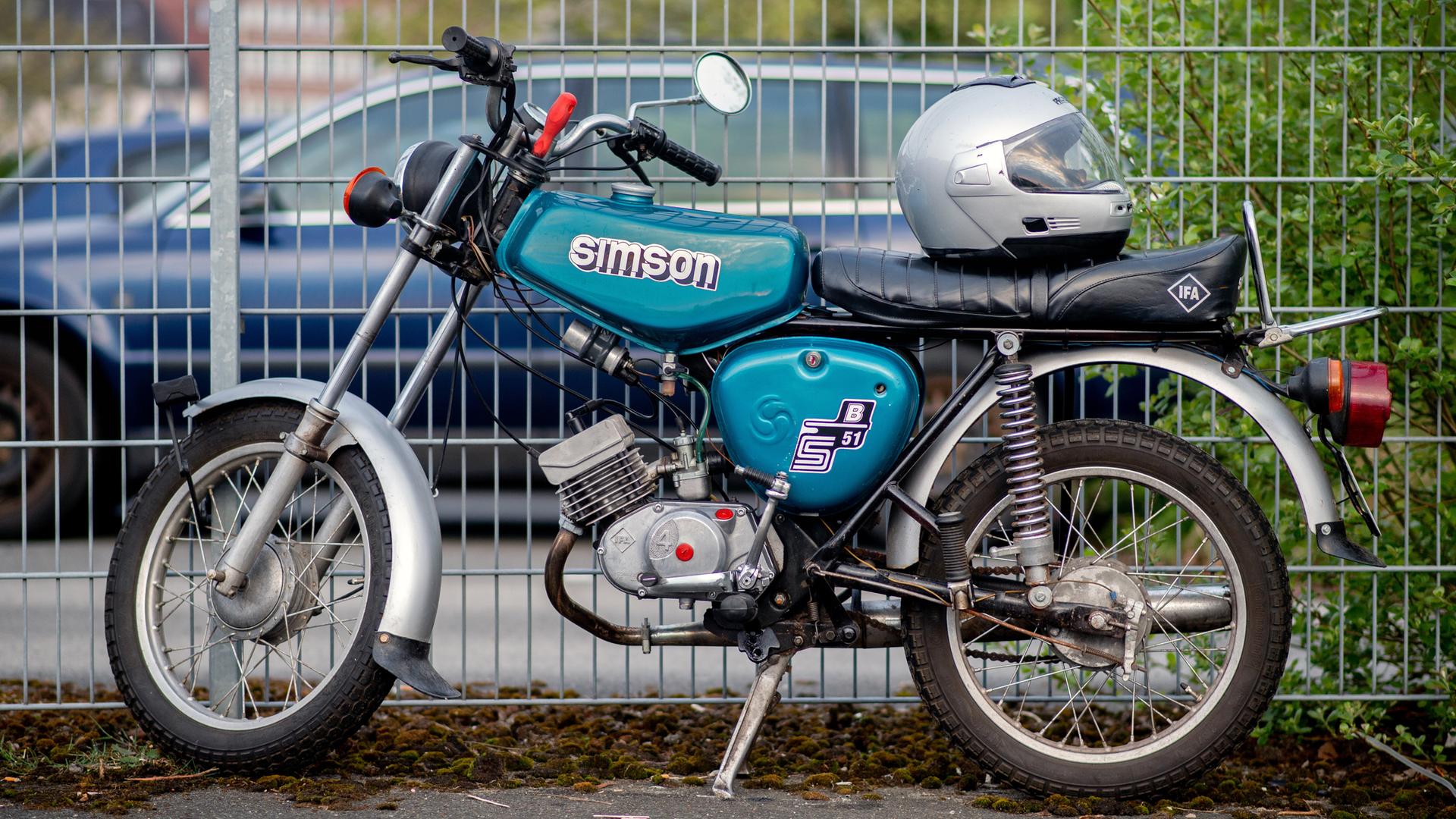 Ein Moped von der Marke Simson steht vor einem Zaun. Auf dem Moped liegt ein Motorrad-Helm.