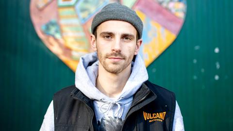 Fynn Kliemann, mit grauer Wollmütze, Kapuzenjacke und schwarzer Weste, steht vor einer grünen Wellblechwand mit einem stilisierten bunten Herz.