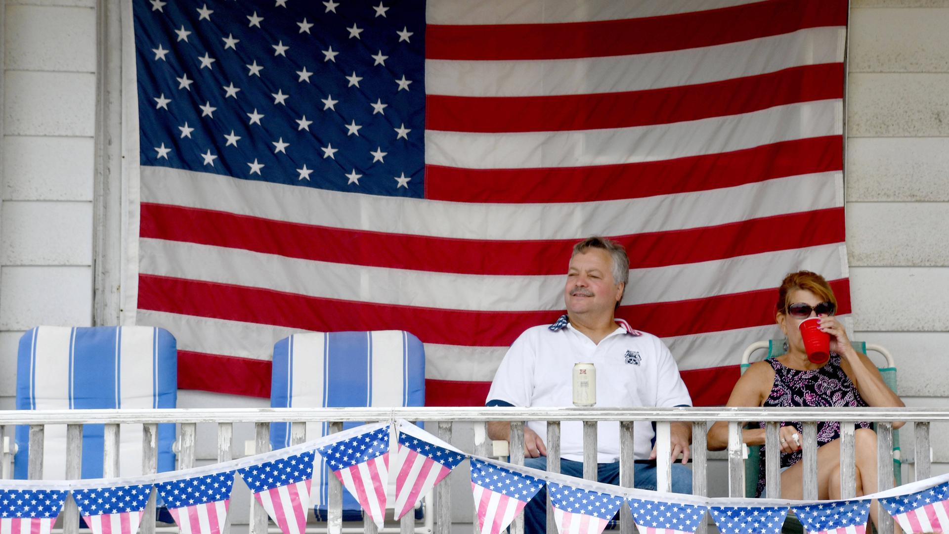 Ein amerikanisches Paar wartet auf den Beginn der Parade zum 4. Juli in Racine, Wisconsin, 2024. Sie sitzen auf ihrer Veranda vor einer riesigen USA-Flagge und trinken aus roten Plastikbechern.