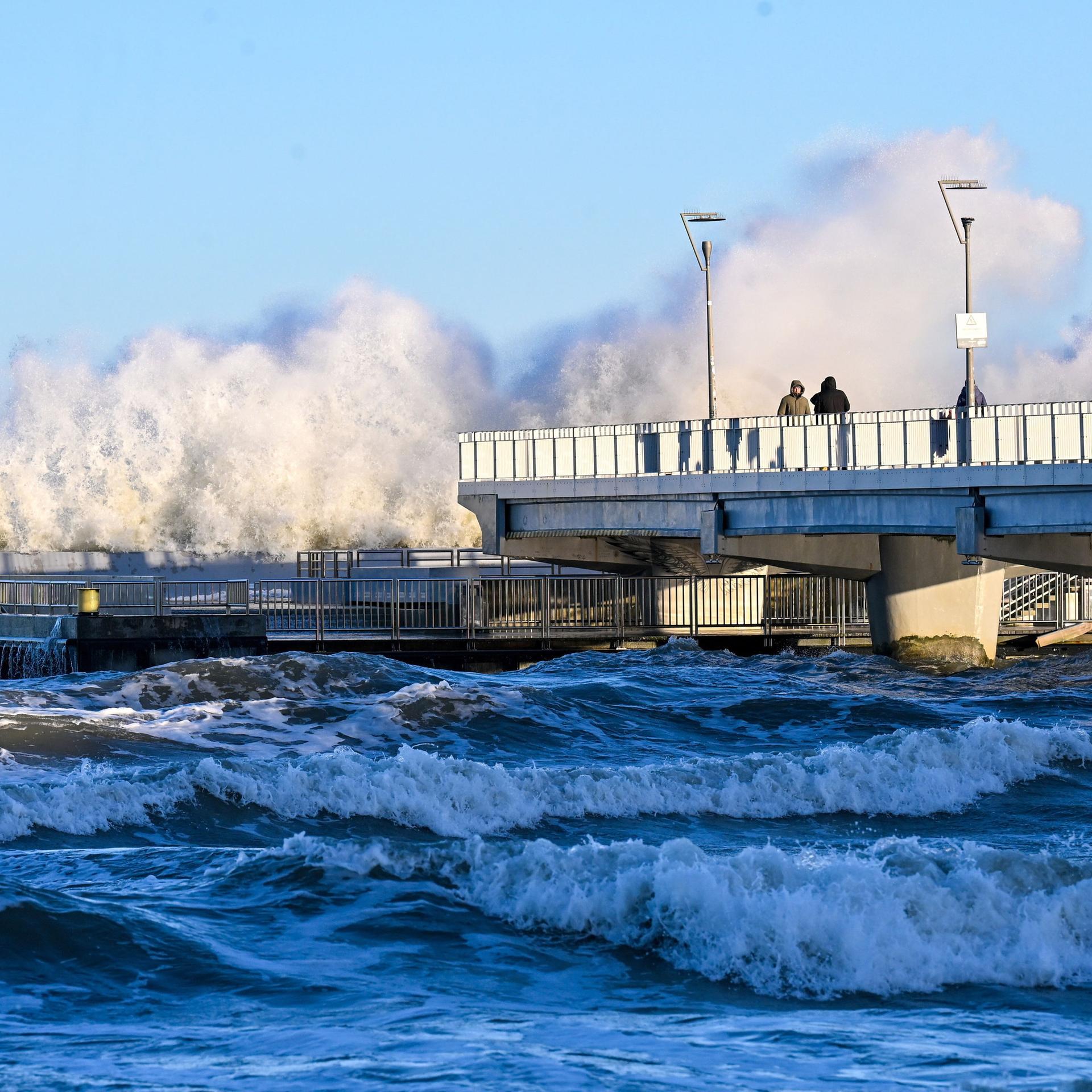 Blick über die aufgewühlte Bucht: Die Wellen schlagen meterhoch über das äußere Ende der Brücke. Am vorderen Ende stehen Menschen und beobachten das Spektakel. 