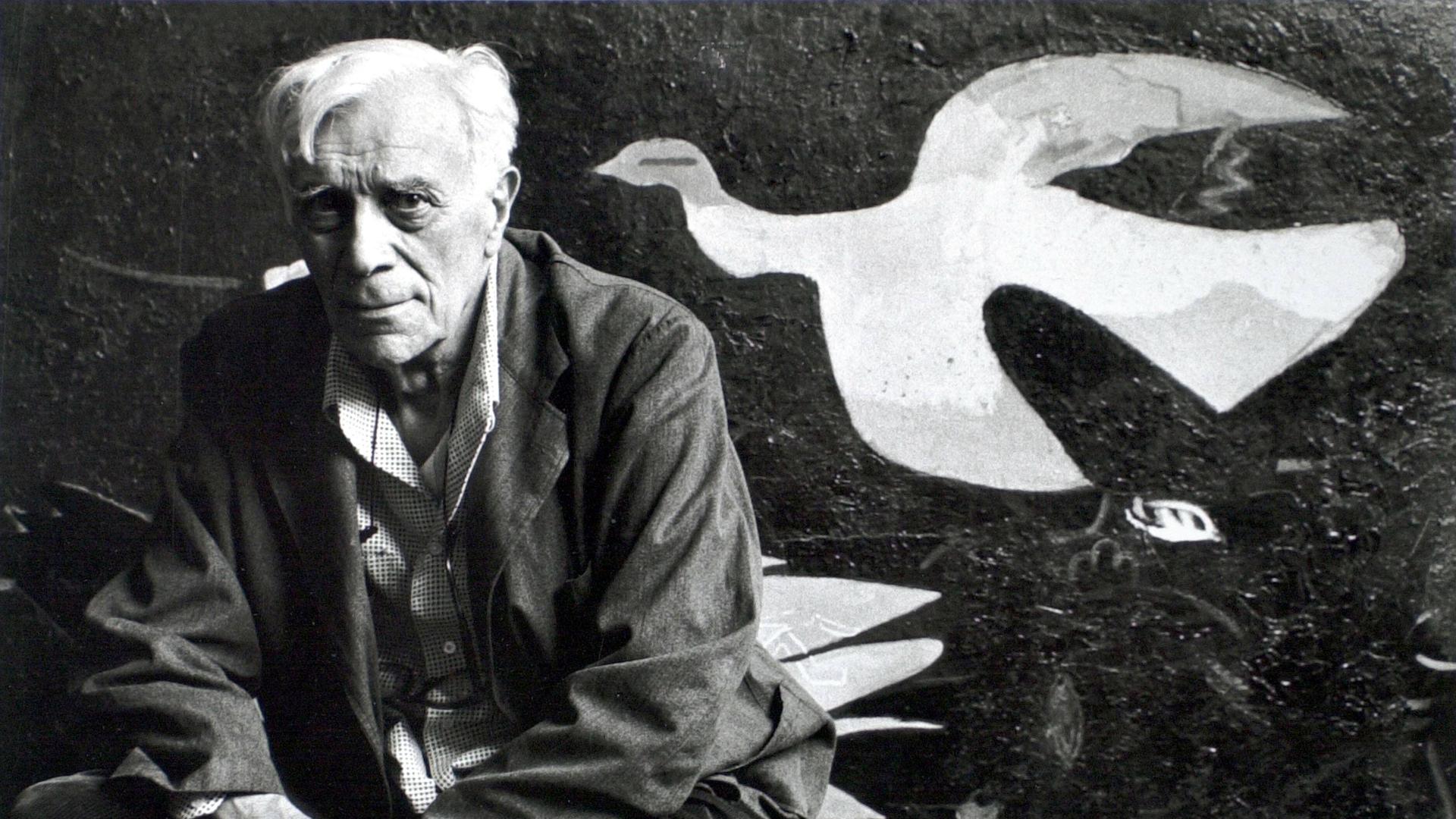 Schwarzweiß-Foto zeigt in die Kamera blickenden Mann vor einer Leinwand mit kubistisch abstrahierter Taube