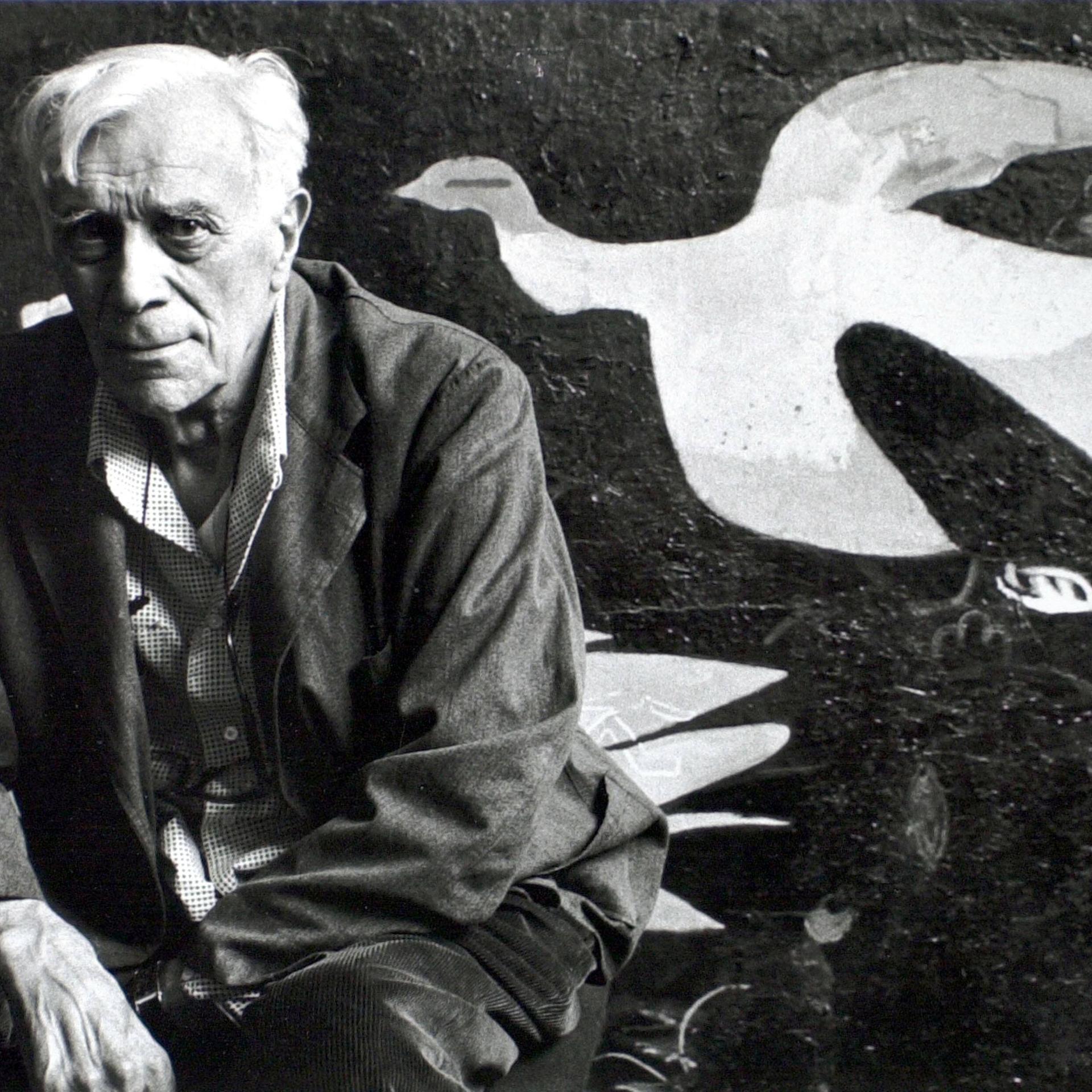 Schwarzweiß-Foto zeigt in die Kamera blickenden Mann vor einer Leinwand mit kubistisch abstrahierter Taube