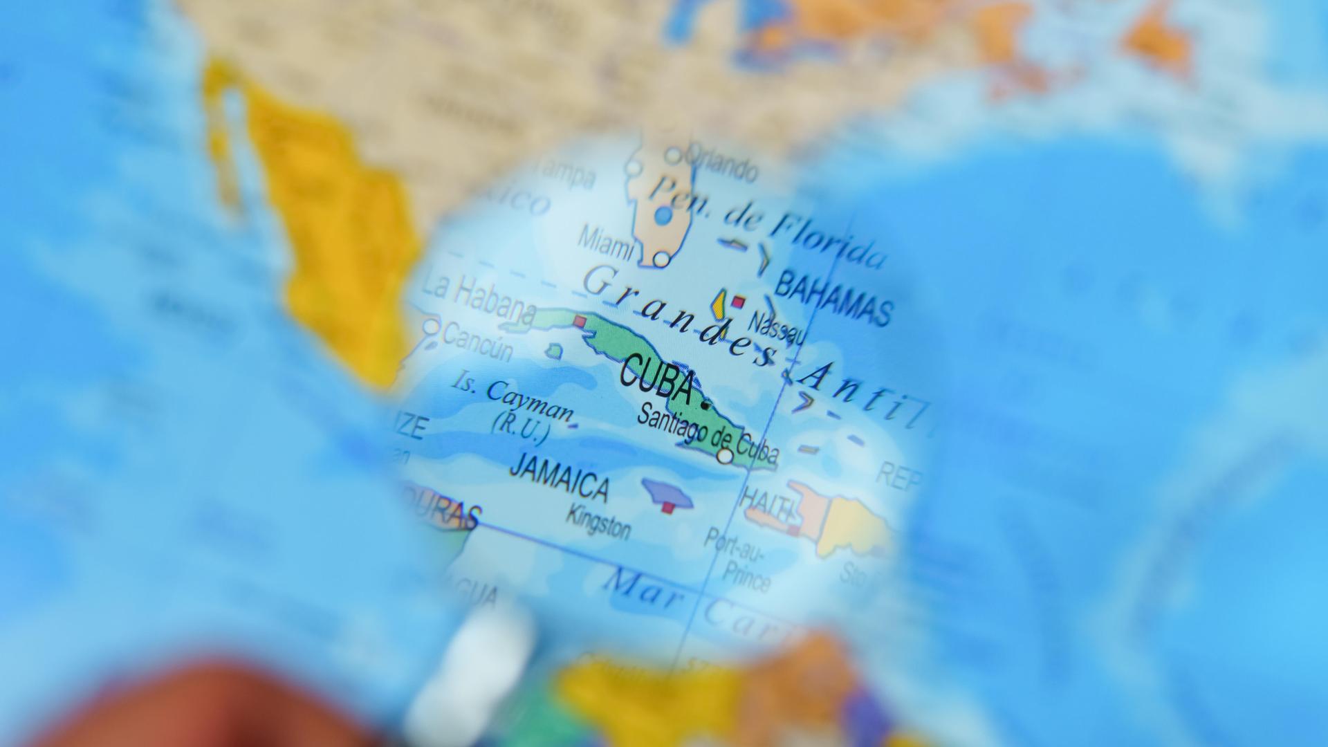 Hand eines Mannes mit einem Vergrößerungsglas, der Kuba auf einer Weltkarte betrachtet.