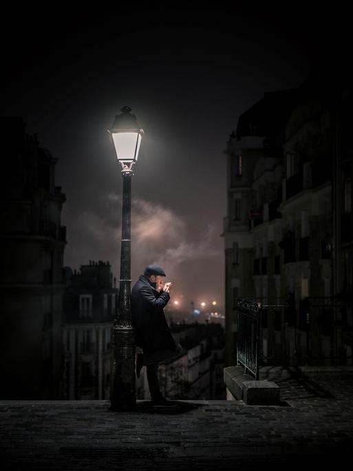 Im nächtlichen Paris lehnt ein Mann an einer Laterne und zündet sich eine Zigarette an.