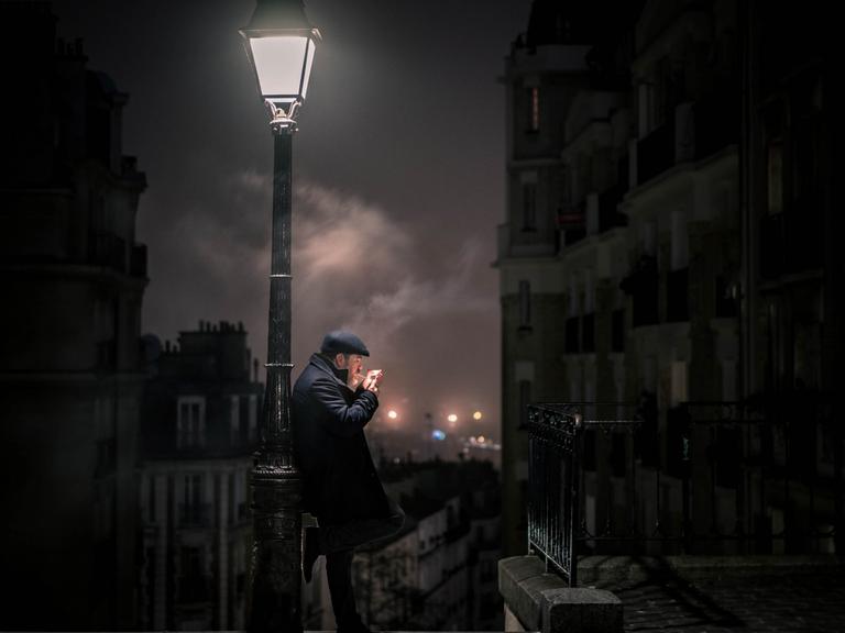 Im nächtlichen Paris lehnt ein Mann an einer Laterne und zündet sich eine Zigarette an.