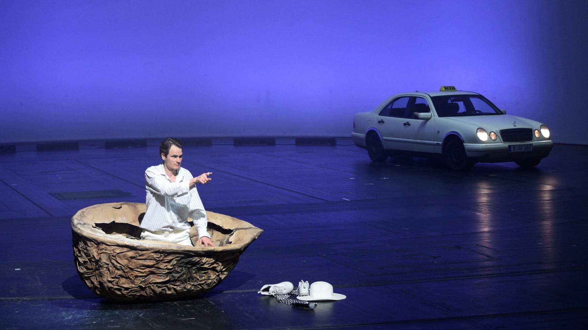 Ein Mann sitzt auf einer Bühne in einer riesigen Nussschale und zeigt in die Ferne. Auf dem Bühnenboden liegen Turnschuhe und ein Hut. Im Hintergrund steht ein Taxi.