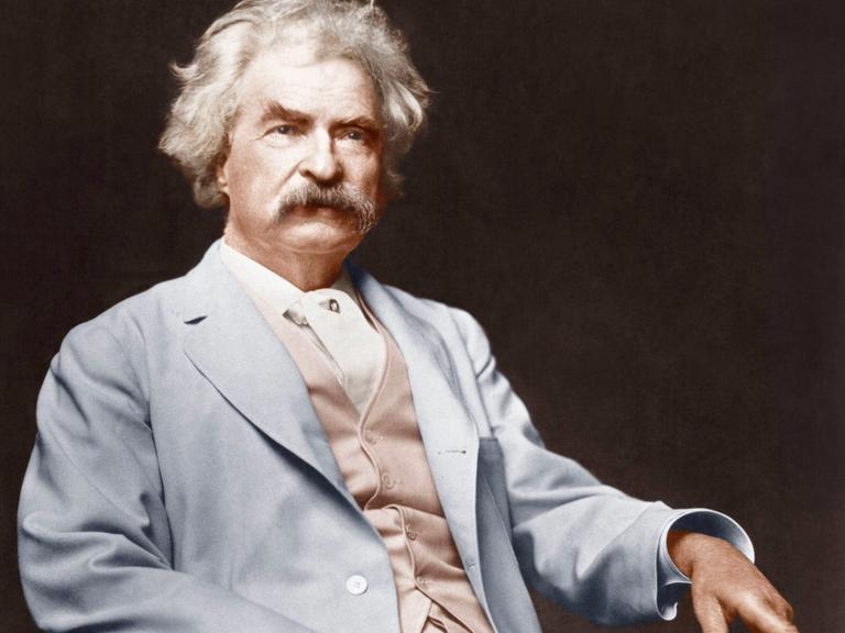 Mark Twain auf einem Foto um 1900. Er sitzt in feinem Zwirn, hat weiße Haare und einen kräftigen Schnauzbart.