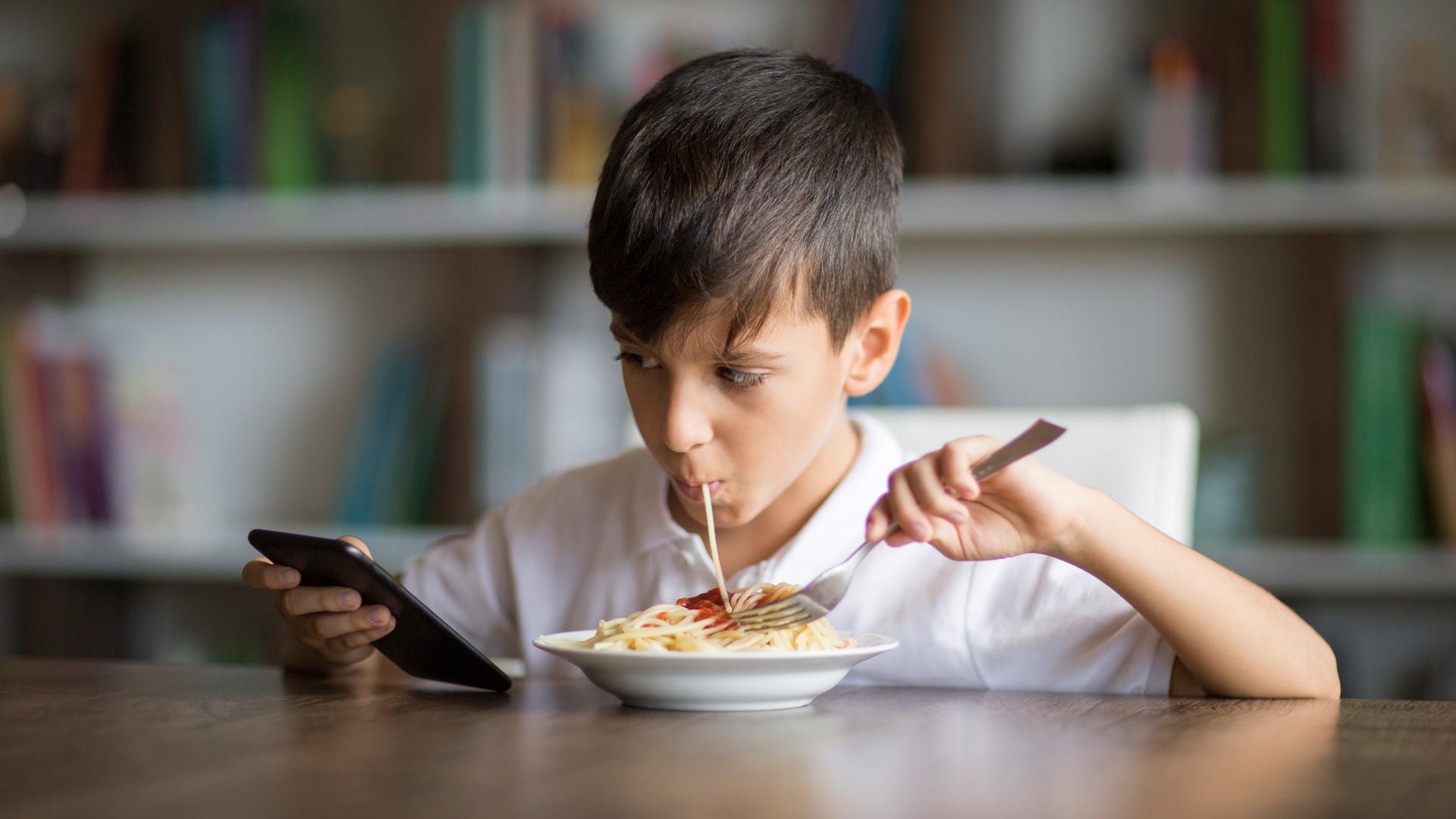 Ein Junge isst Spaghetti und schaut dabei auf das Handy in seiner Hand