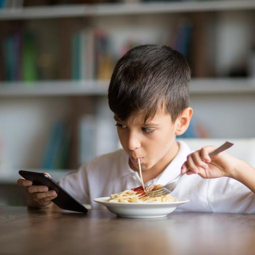 Ein Junge isst Spaghetti und schaut dabei auf das Handy in seiner Hand