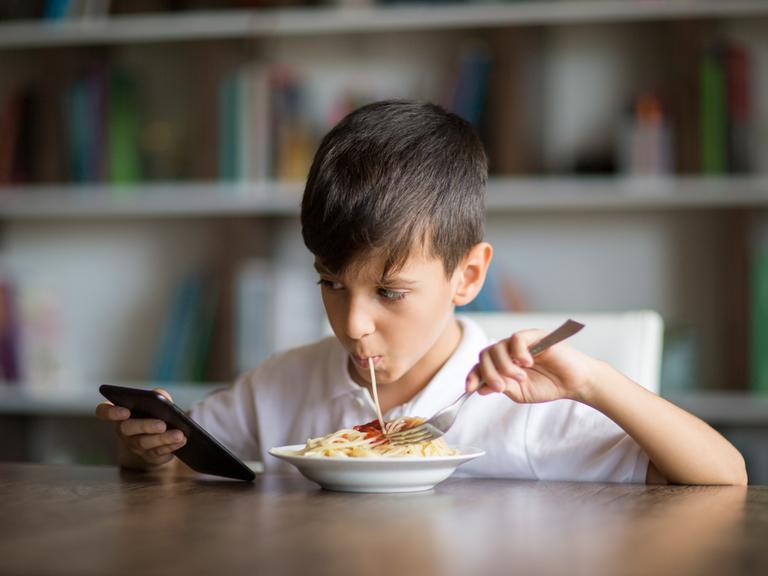 Ein Junge isst Spaghetti und schaut dabei auf das Handy in seiner Hand
