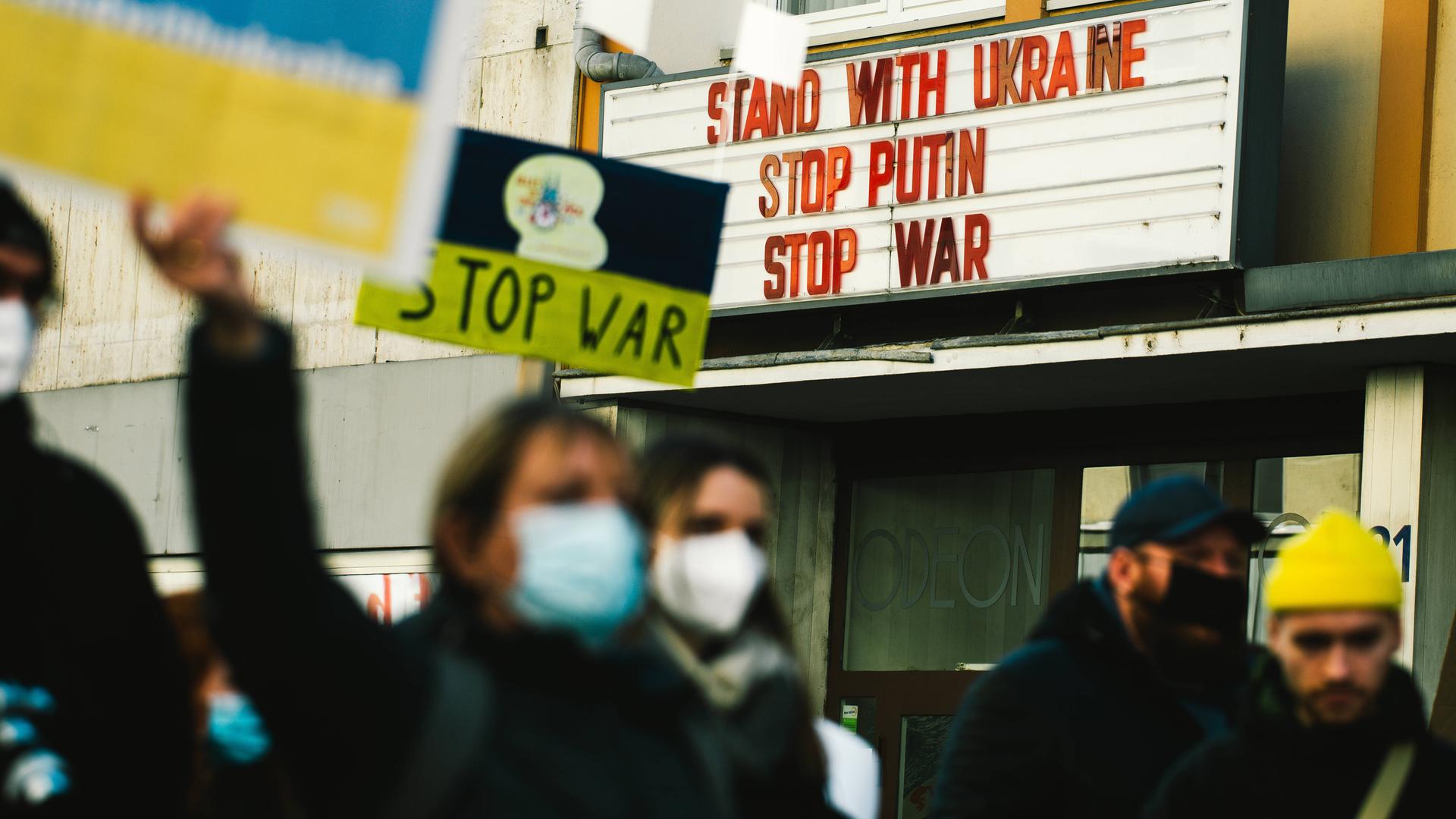 Demonstranten gehen an einem Kino vorbei. An dessen Hausfassade steht: Solidarität mit der Ukraine. Stoppt Putin. Stoppt den Krieg.
