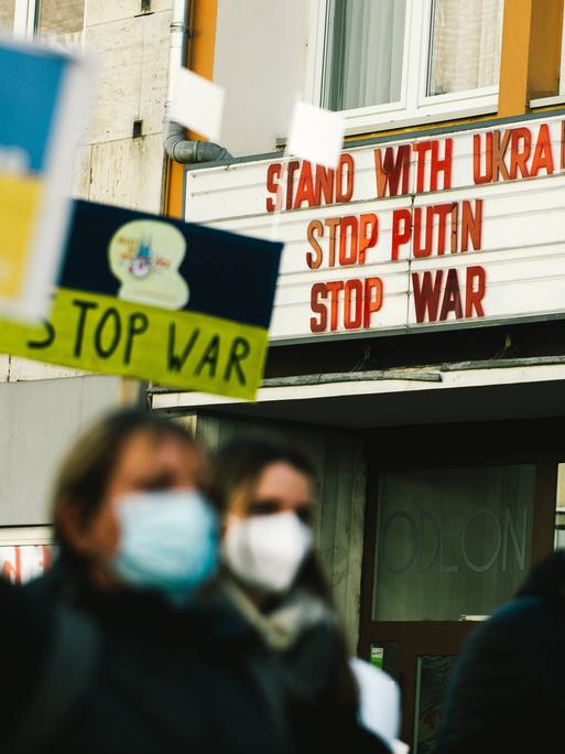 Demonstranten gehen an einem Kino vorbei. An dessen Hausfassade steht: Solidarität mit der Ukraine. Stoppt Putin. Stoppt den Krieg.