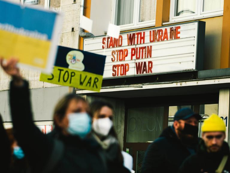 Demonstranten gehen an einem Kino vorbei. An dessen Hausfassade steht: Solidarität mit der Ukraine. Stoppt Putin. Stoppt den Krieg.