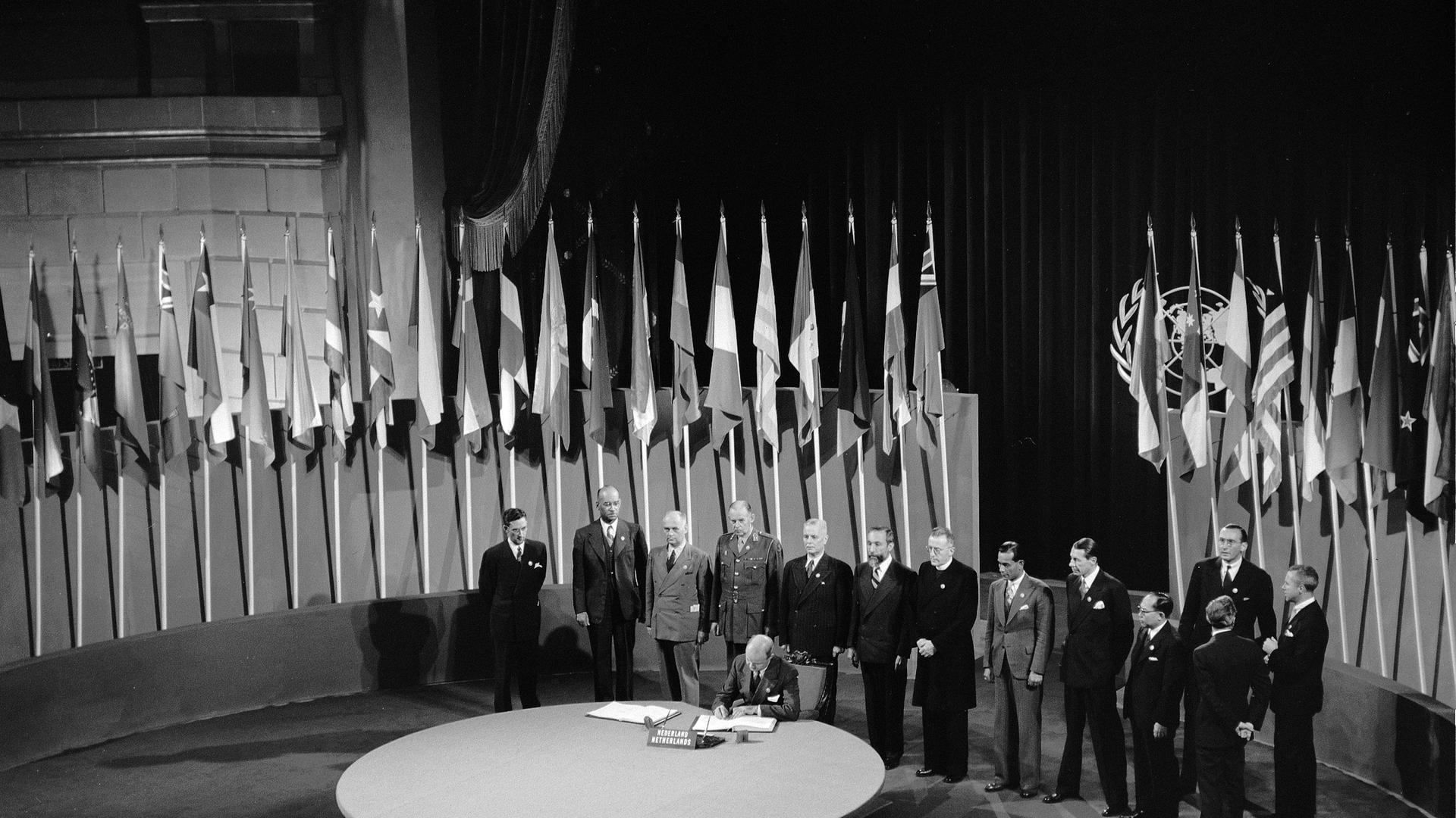 Schwarz-weiss Foto: An einem runden Tisch vor vielen Flaggen und mit zahlreichen Personen im Hintergrund unterschreibt der niederländische Botschafter in den USA die Charta der Vereinten Nationen.