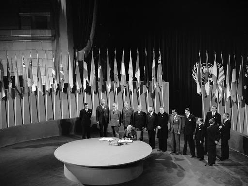 Schwarz-weiss Foto: An einem runden Tisch vor vielen Flaggen und mit zahlreichen Personen im Hintergrund unterschreibt der niederländische Botschafter in den USA die Charta der Vereinten Nationen.
