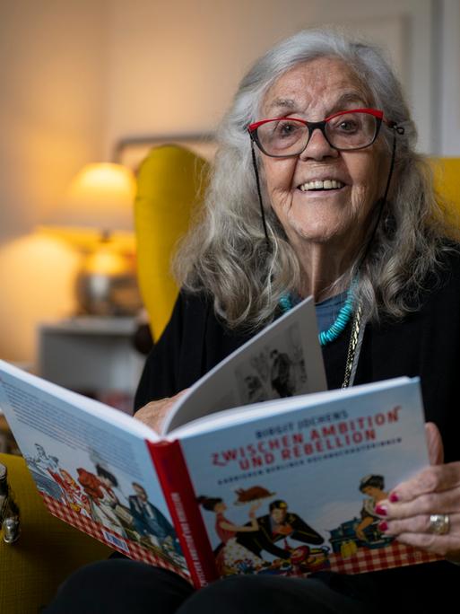 Ursula Winnington, Kochbuchautorin und Köchin, sitzt in einem Sessel und hält ein Kochbuch in den Händen. 