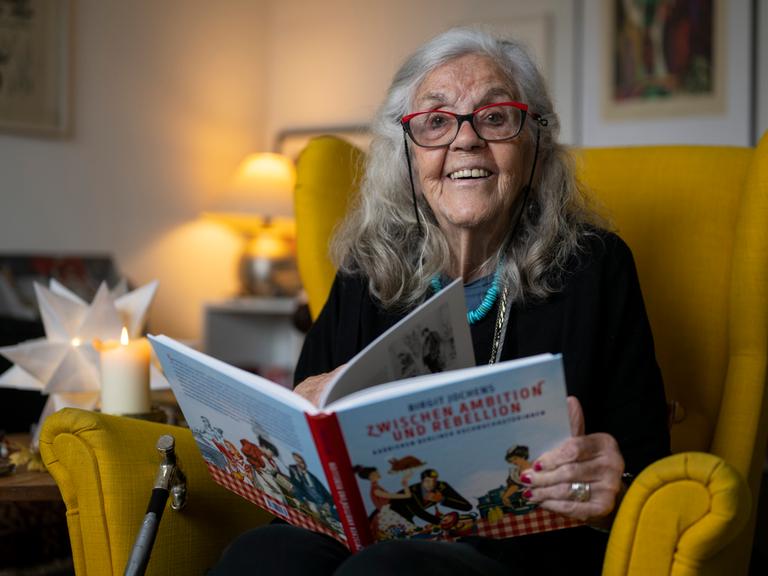 Ursula Winnington, Kochbuchautorin und Köchin, sitzt in einem Sessel und hält ein Kochbuch in den Händen. 