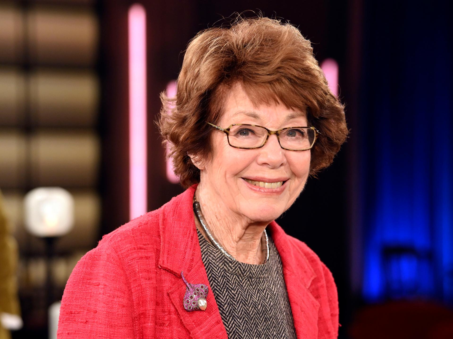 Marianne Koch steht 2025 in einem Fernsehstudio, trägt einen roten Blazer und ein Oberteil in Fischgrätmuster und lächelt.