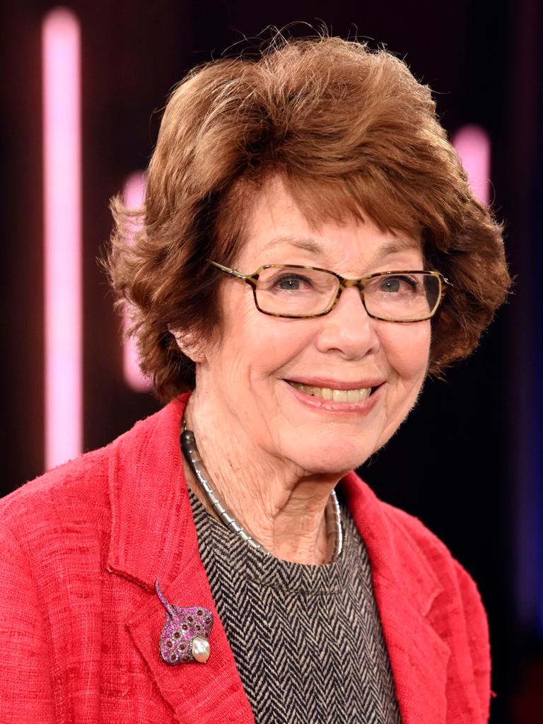 Marianne Koch steht 2025 in einem Fernsehstudio, trägt einen roten Blazer und ein Oberteil in Fischgrätmuster und lächelt.