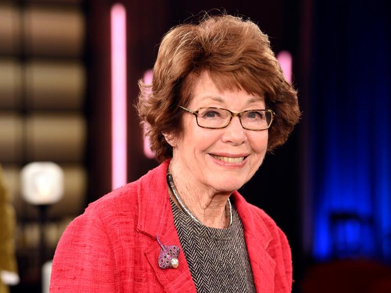 Marianne Koch steht 2025 in einem Fernsehstudio, trägt einen roten Blazer und ein Oberteil in Fischgrätmuster und lächelt.