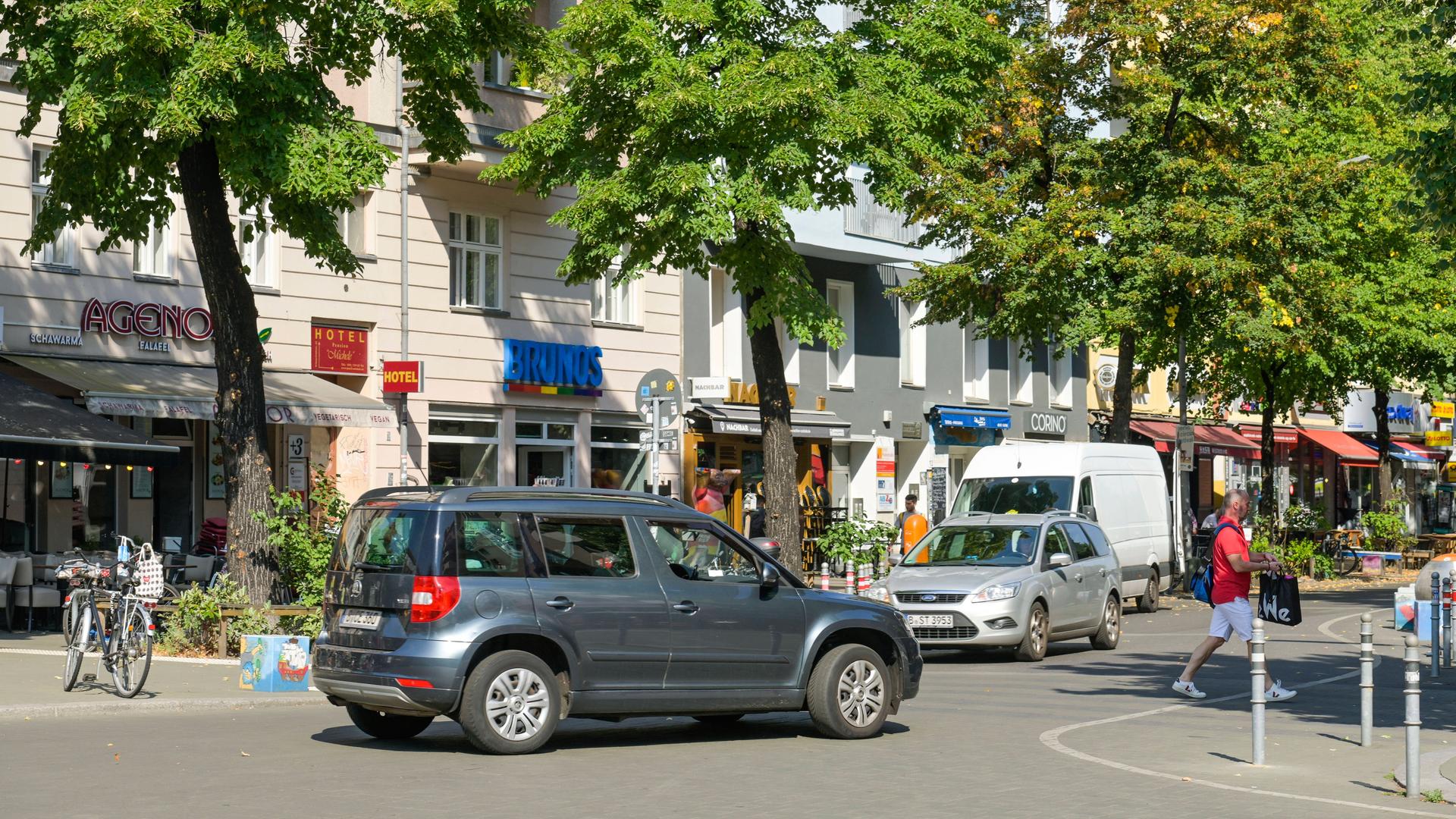 Auf einer Straße fahren zwei Autos. Ein Fußgänger geht über die Straße. Am Rand der Straße steht ein Fahrrad. An der Seite sind Bäume und Geschäfte. Die Sonne scheint.