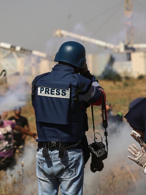 Ein Mann mit einer kugelsicheren Weste, die die Aufschrift „PRESS“ trägt, steht mit dem Rücken zur Kamera vor einem verdorrten Feld, aus dem vereinzelt Rauch aufsteigt. 