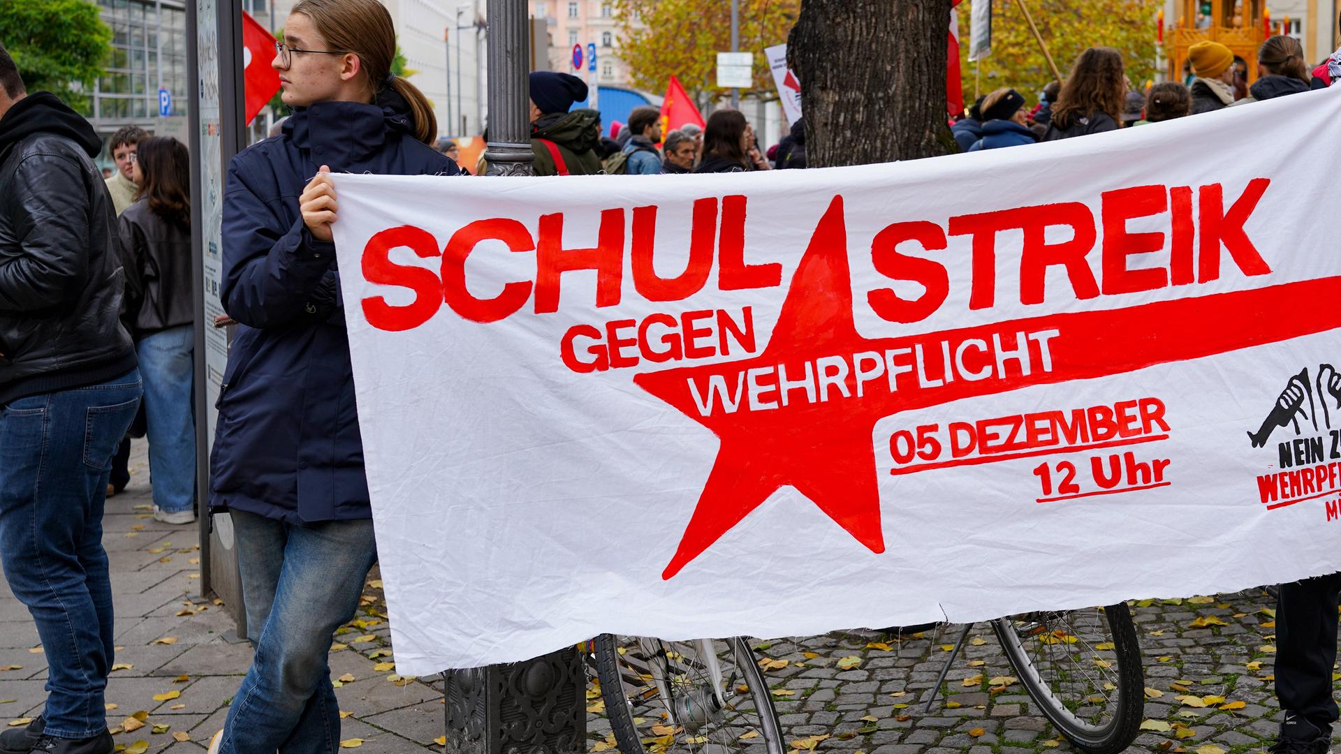 Ein junger Mann hält ein Plakat fest, darauf steht: Schulstreik gegen Wehrpflicht, 5. Dezember, 12 Uhr. Das Foto ist von einer Demo in München, vom 8. November 2025.