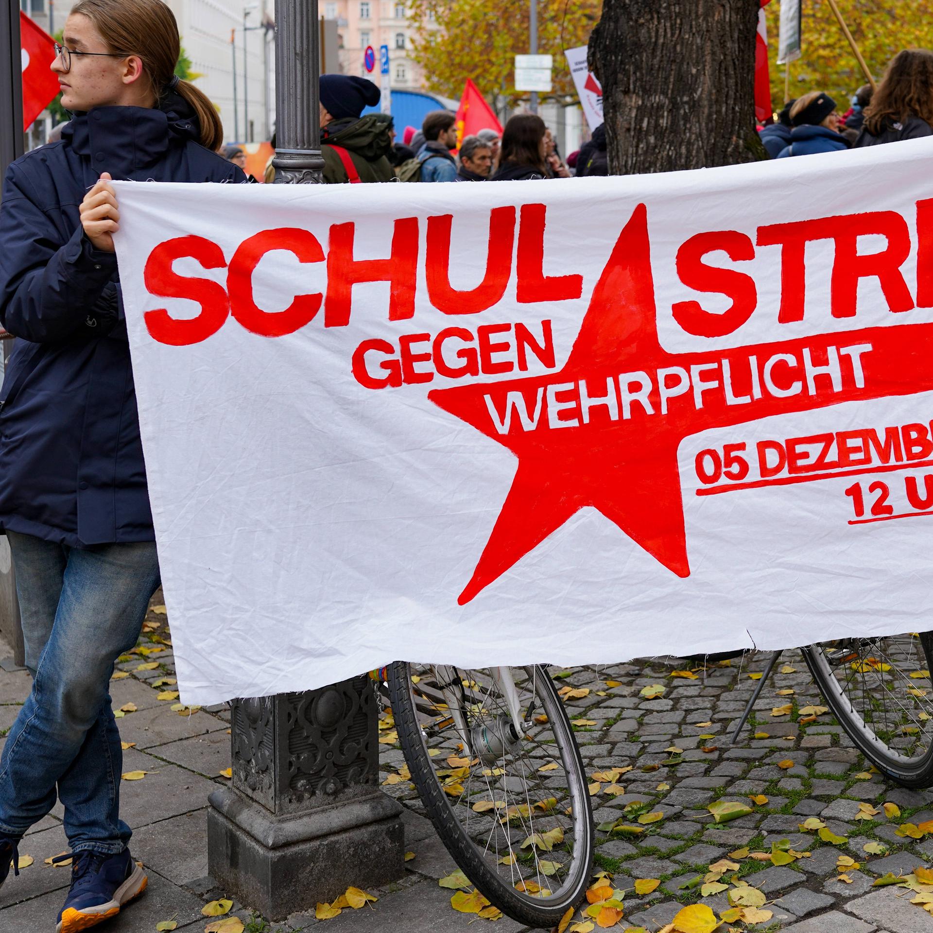 Ein junger Mann hält ein Plakat fest, darauf steht: Schulstreik gegen Wehrpflicht, 5. Dezember, 12 Uhr. Das Foto ist von einer Demo in München, vom 8. November 2025.