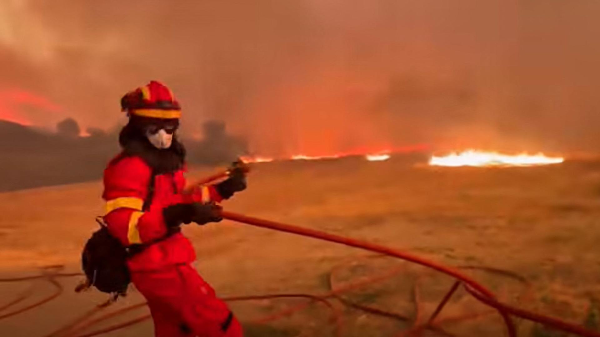 Der Mann ist gekleidet wie ein Feuerwehrmann. Er zieht einen Schlauch über ein Feld. Im Hintergrund Flammen und Rauch. Die Szenerie ist in rotes Licht gekleidet. 