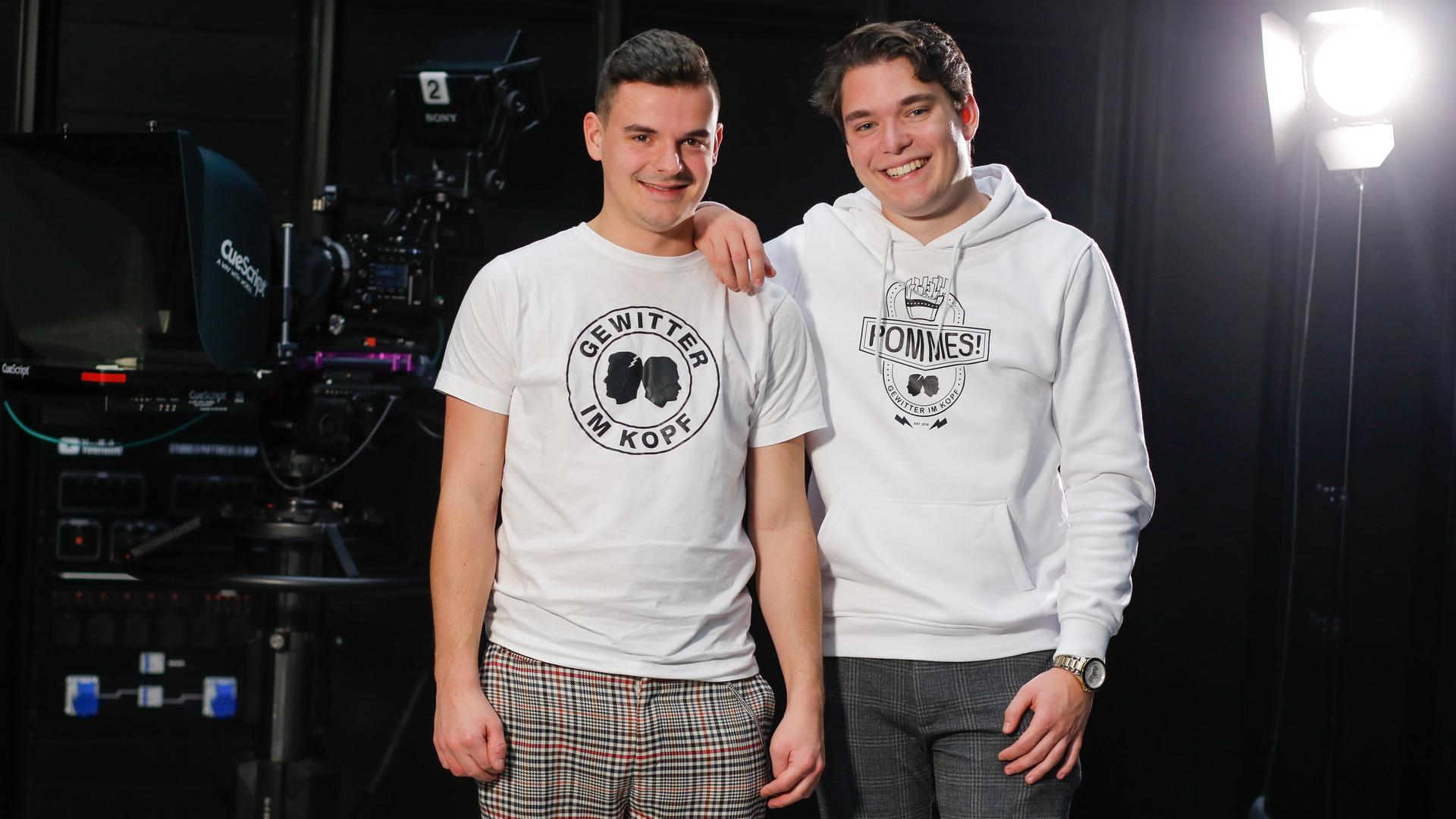 Jan Zimmermann (links) trägt ein T-Shirt mit der Aufschrift "Gewitter im Kopf" und steht mit seinem Kompagnon Tim Lehmann lachend auf einer Bühne. 
