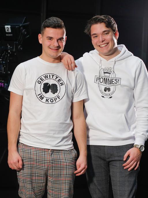 Jan Zimmermann (links) trägt ein T-Shirt mit der Aufschrift "Gewitter im Kopf" und steht mit seinem Kompagnon Tim Lehmann lachend auf einer Bühne. 