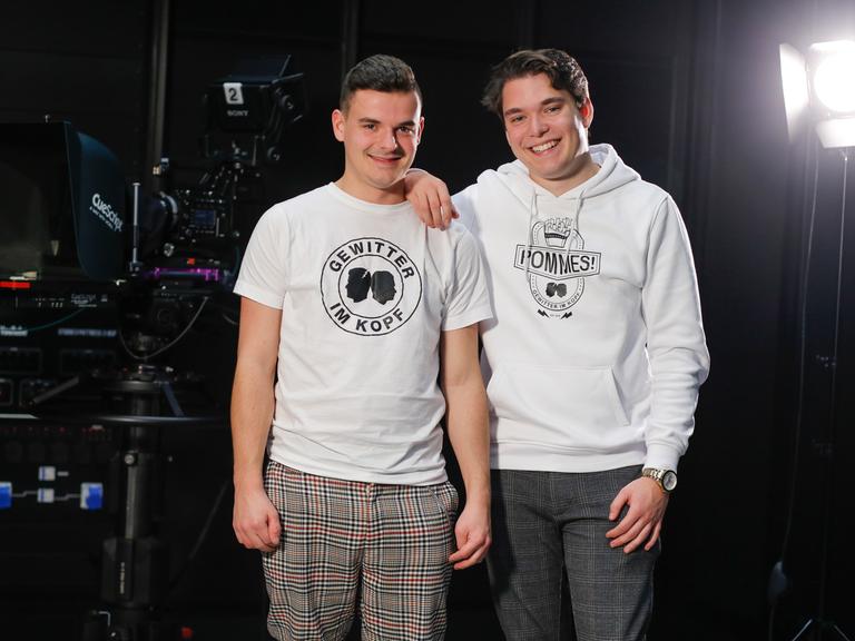 Jan Zimmermann (links) trägt ein T-Shirt mit der Aufschrift "Gewitter im Kopf" und steht mit seinem Kompagnon Tim Lehmann lachend auf einer Bühne. 