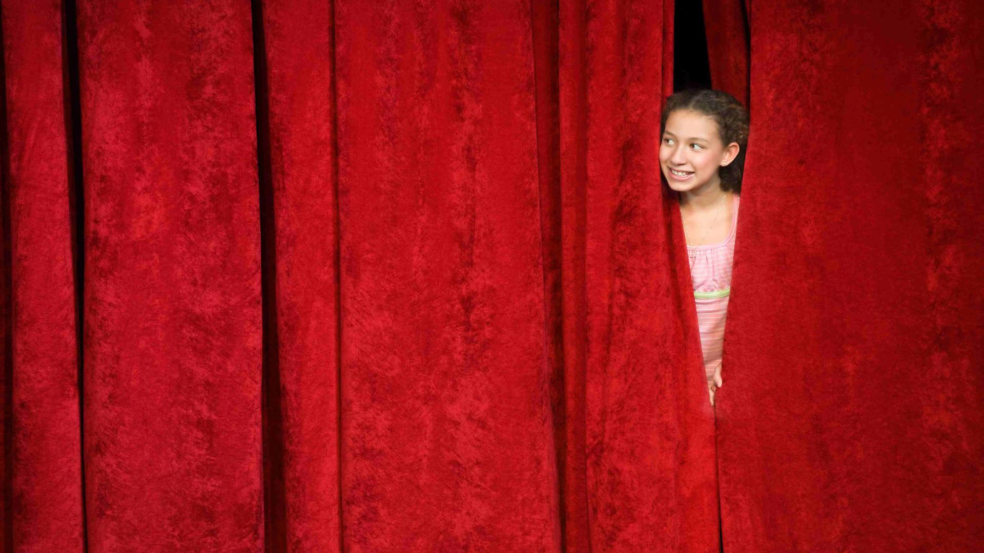 Ein Mädchen spickt von hinter der Bühne durch den geschlossenen roten Vorhang nach draußen.