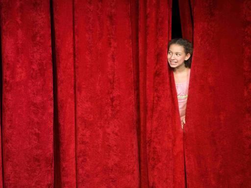 Ein Mädchen spickt von hinter der Bühne durch den geschlossenen roten Vorhang nach draußen.