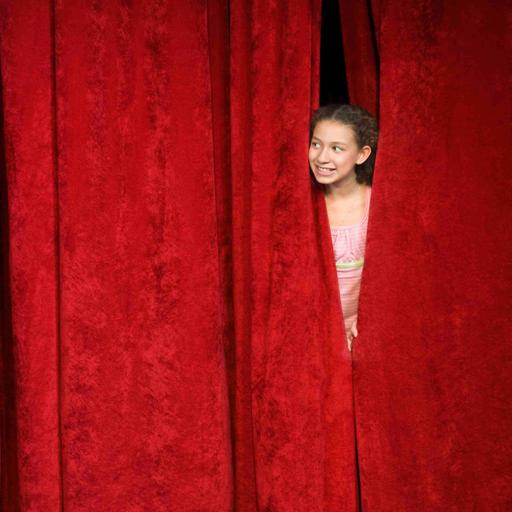 Ein Mädchen spickt von hinter der Bühne durch den geschlossenen roten Vorhang nach draußen.