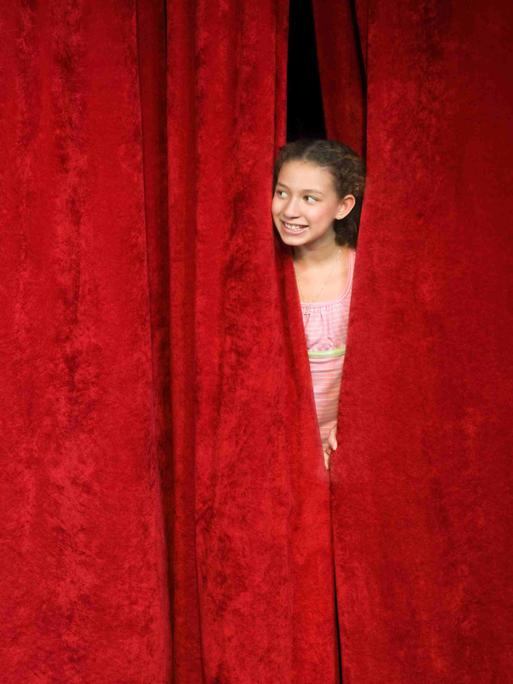 Ein Mädchen spickt von hinter der Bühne durch den geschlossenen roten Vorhang nach draußen.