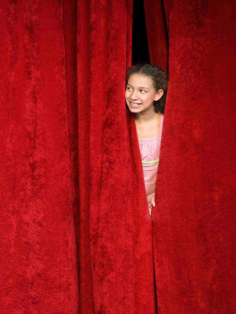 Ein Mädchen spickt von hinter der Bühne durch den geschlossenen roten Vorhang nach draußen.