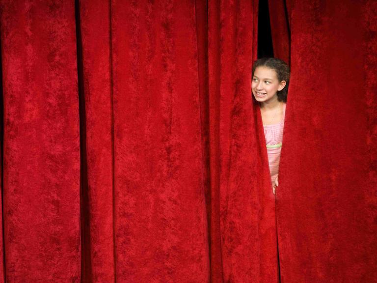 Ein Mädchen spickt von hinter der Bühne durch den geschlossenen roten Vorhang nach draußen. Ein Mädchen spickt von hinter der Bühne durch den geschlossenen roten Vorhang nach draußen.