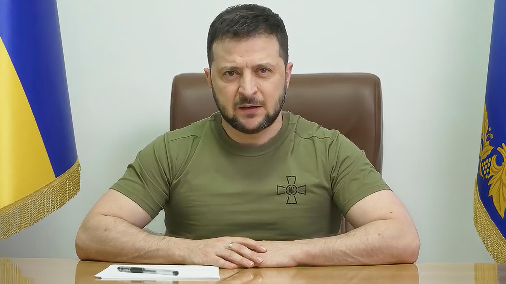 Selenskyj im olivgrünen T-Shirt sitzt an einem Tisch und spricht. Rechts und links von ihm jeweils eine ukrainische Fahne.