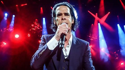 Nick Cave steht im Scheinwerferlicht auf der Bühne und singt frontal in die Kamera