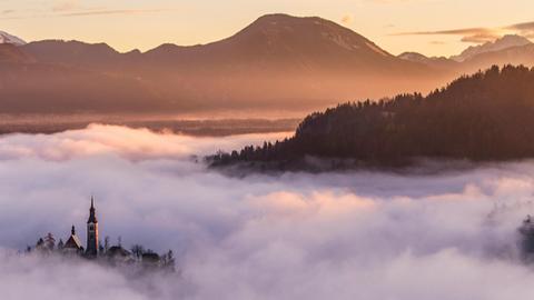 Blick von einem Berg in ein Tal, das voller Nebel ist, während wenige Gebäude hoch genug sind, um wie eine Insel hervorzulugen.