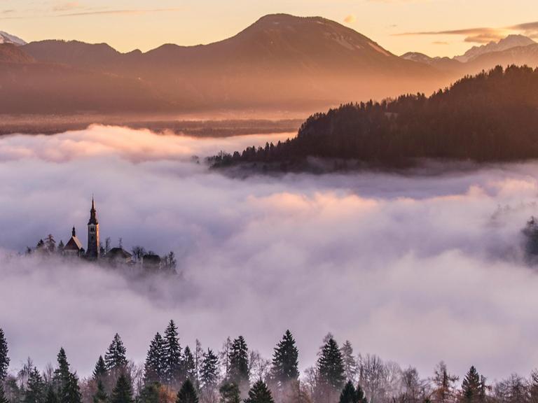 Blick von einem Berg in ein Tal, das voller Nebel ist, während wenige Gebäude hoch genug sind, um wie eine Insel hervorzulugen.