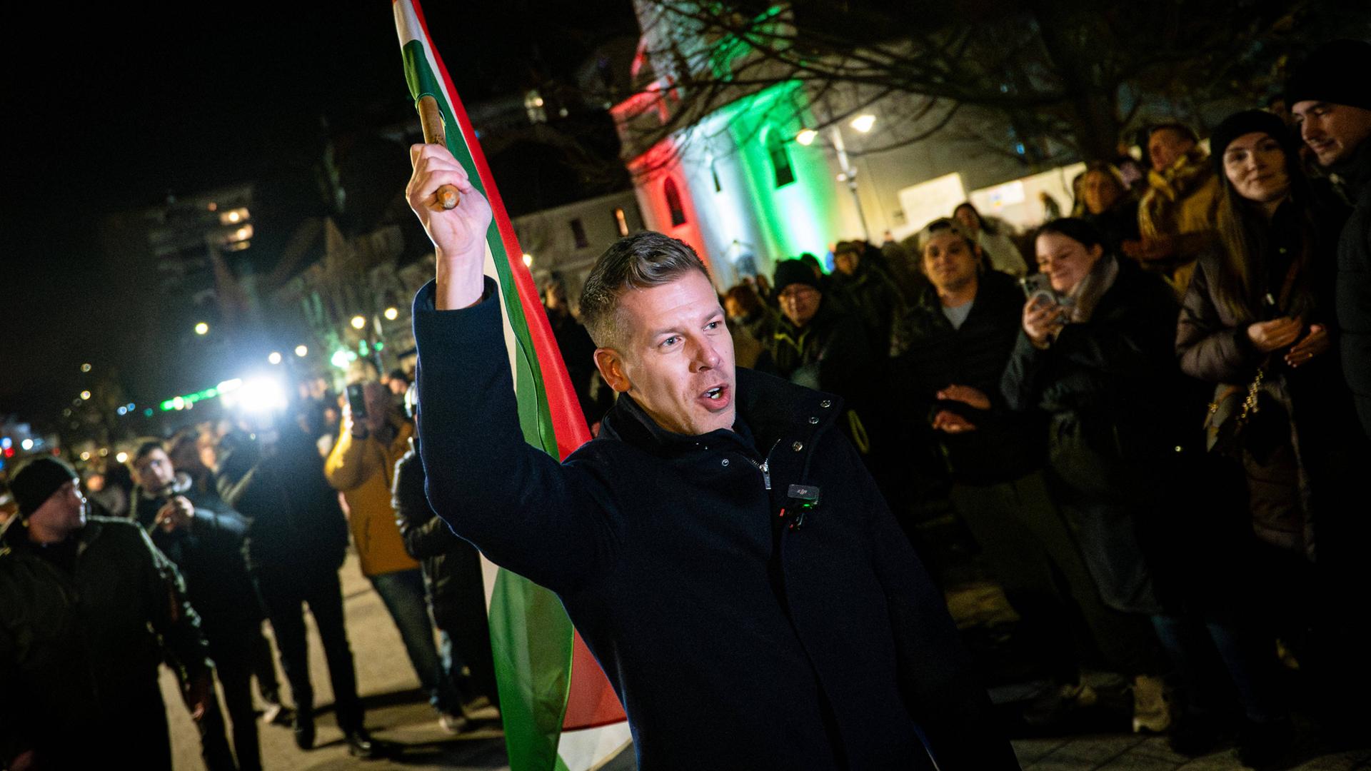 Péter Magyar schwenkt am späten Abend auf einem öffentlichen Platz die ungarische Fahne. Er trägt warme Kleidung.