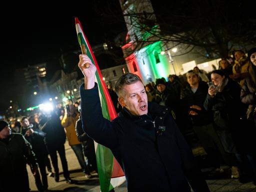Péter Magyar schwenkt am späten Abend auf einem öffentlichen Platz die ungarische Fahne. Er trägt warme Kleidung.