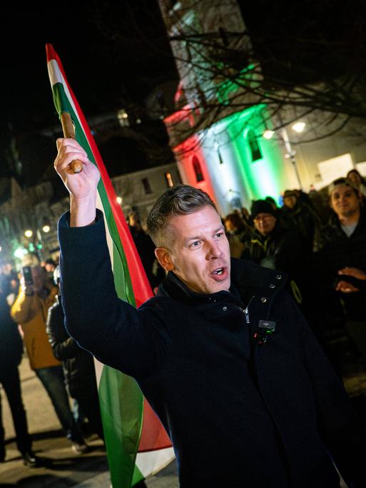 Péter Magyar schwenkt am späten Abend auf einem öffentlichen Platz die ungarische Fahne. Er trägt warme Kleidung.