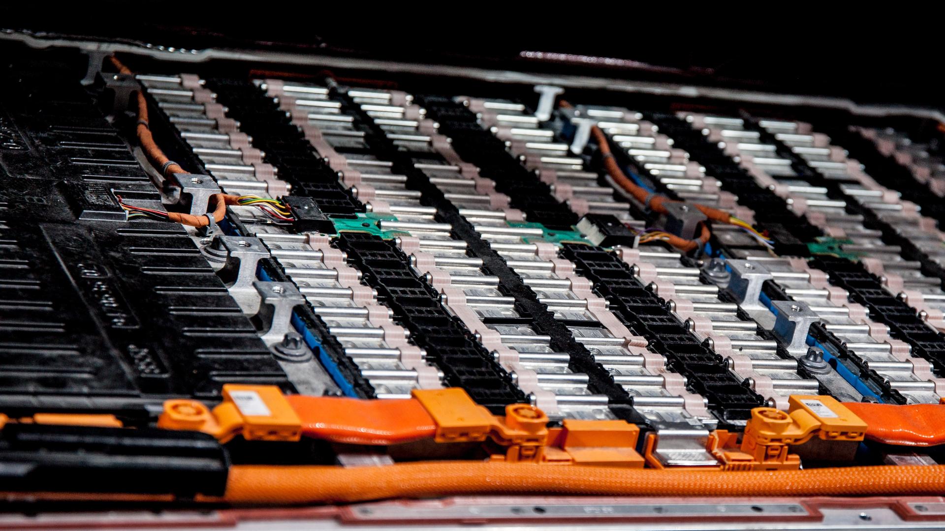 Blick auf die zahlreichen Batterien, die nebeneinander angereiht sind vor einem dunklen Hintergrund. Im Vordergrund orangefarbene Kabel. Blick auf die zahlreichen Batterien, die nebeneinander angereiht sind vor einem dunklen Hintergrund. Im Vordergrund orangefarbene Kabel.