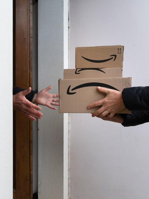 Drei Pakete mit dem Amazon Logo auf der Seite werden an einer Haustür überreicht.